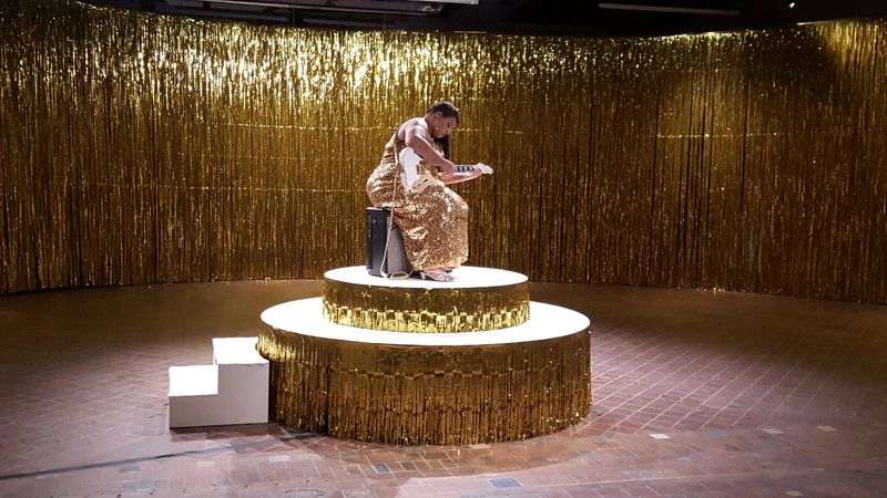 A room is lined with floor to ceiling shimming, golden curtains of streamers. In the middle of the room a circular platform, also draped on it's edges in the gold streams, holds a live woman, clad in a shimmery golden sequin dress. She sits on a speaker, leaned over a white guitar she is preparing to play.