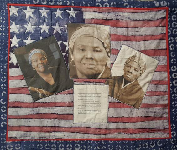 A small quilt hangs on a wall. It is an American flag that has been overlaid with three sepia tone photos of a middle aged African American woman. Below the center photo, we can see a letter that details thoughts about Harriet Tubman.