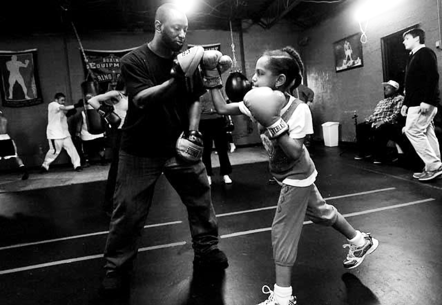 Khali Sweeney stands in the gym, helping a young female child boxer work her gloves. There are boxers working on equipment in the background.