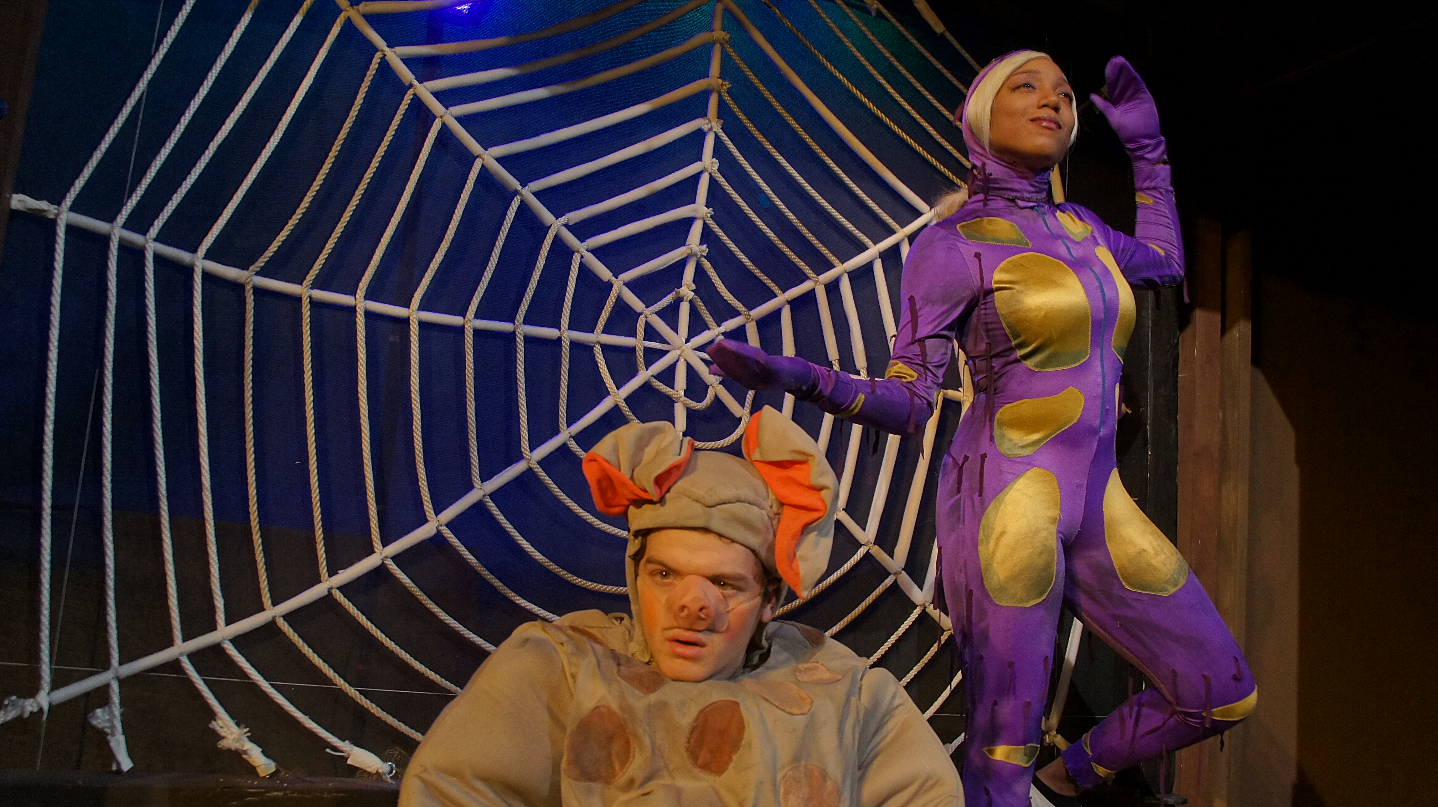 Photo of stage production of Charlotte's Web with large web and actor Dakota Nuttall in Wilbur the pig costume in front of actress Natasha Hawkins in Charlotte the spider costume.