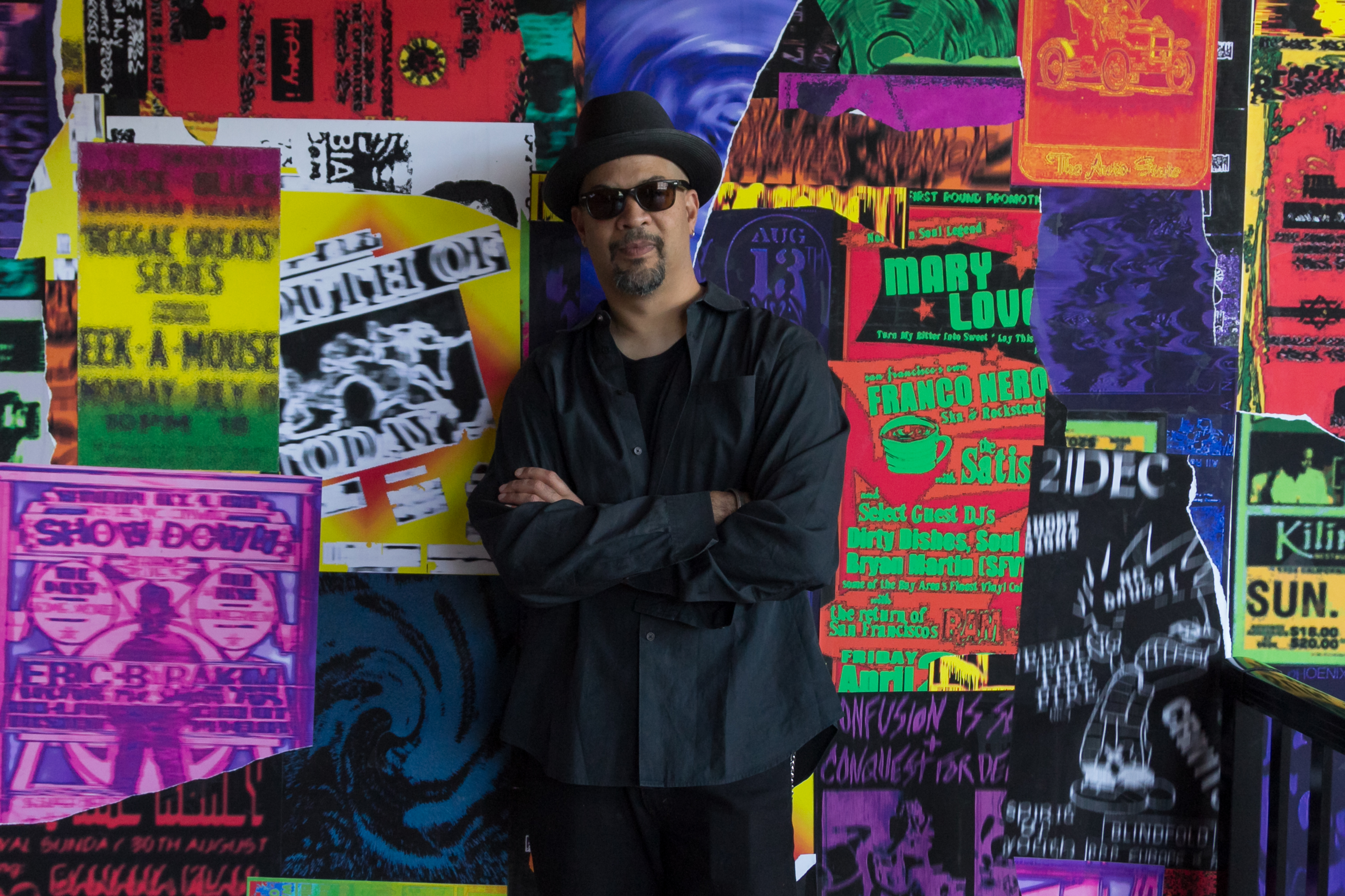 Photo of the artist Gary Simmons in front of his wall of colorful fly posters each with a Detroit theme.