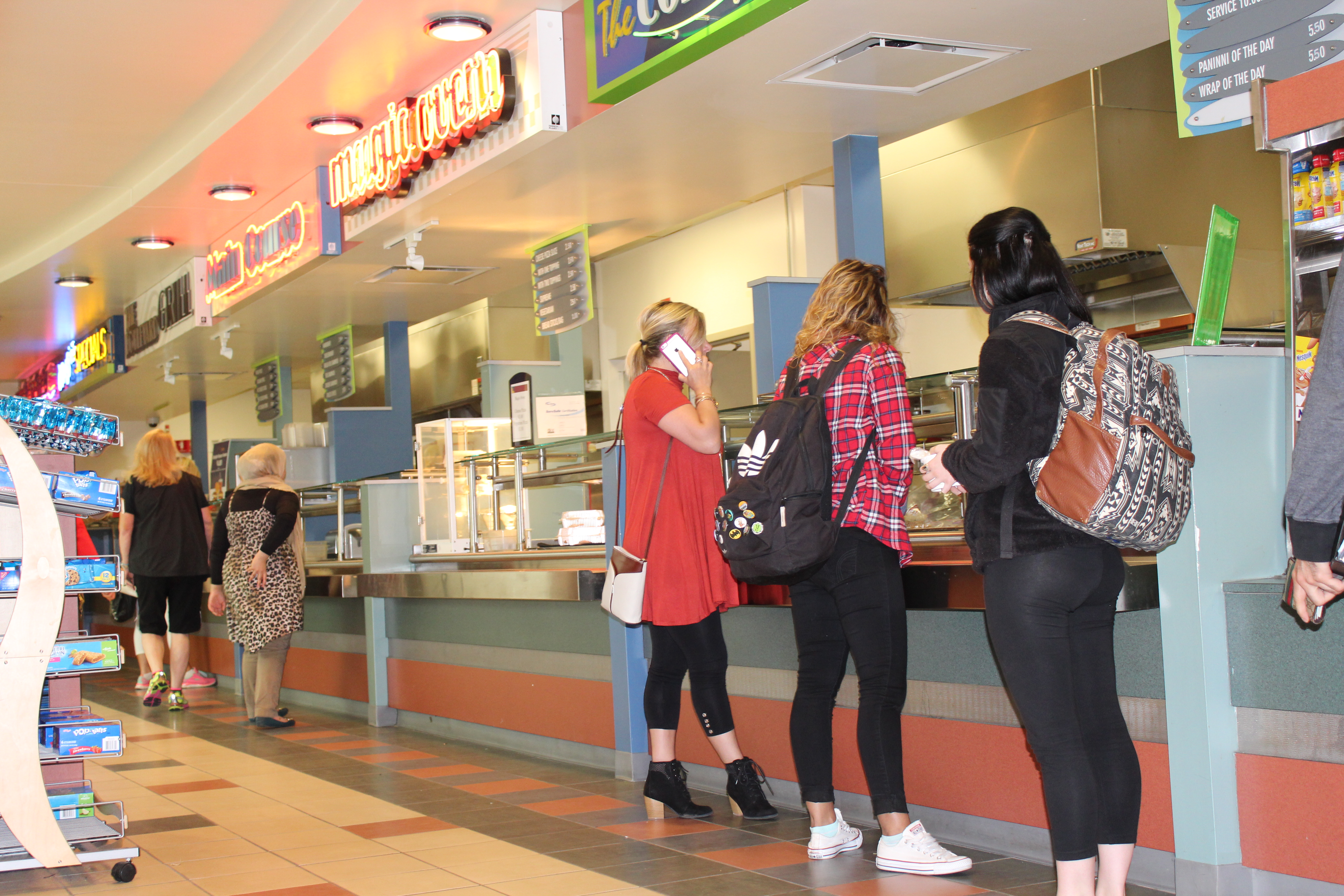 Students waiting for food at the cafeteria serving area with neon signs above them indicating different food options.