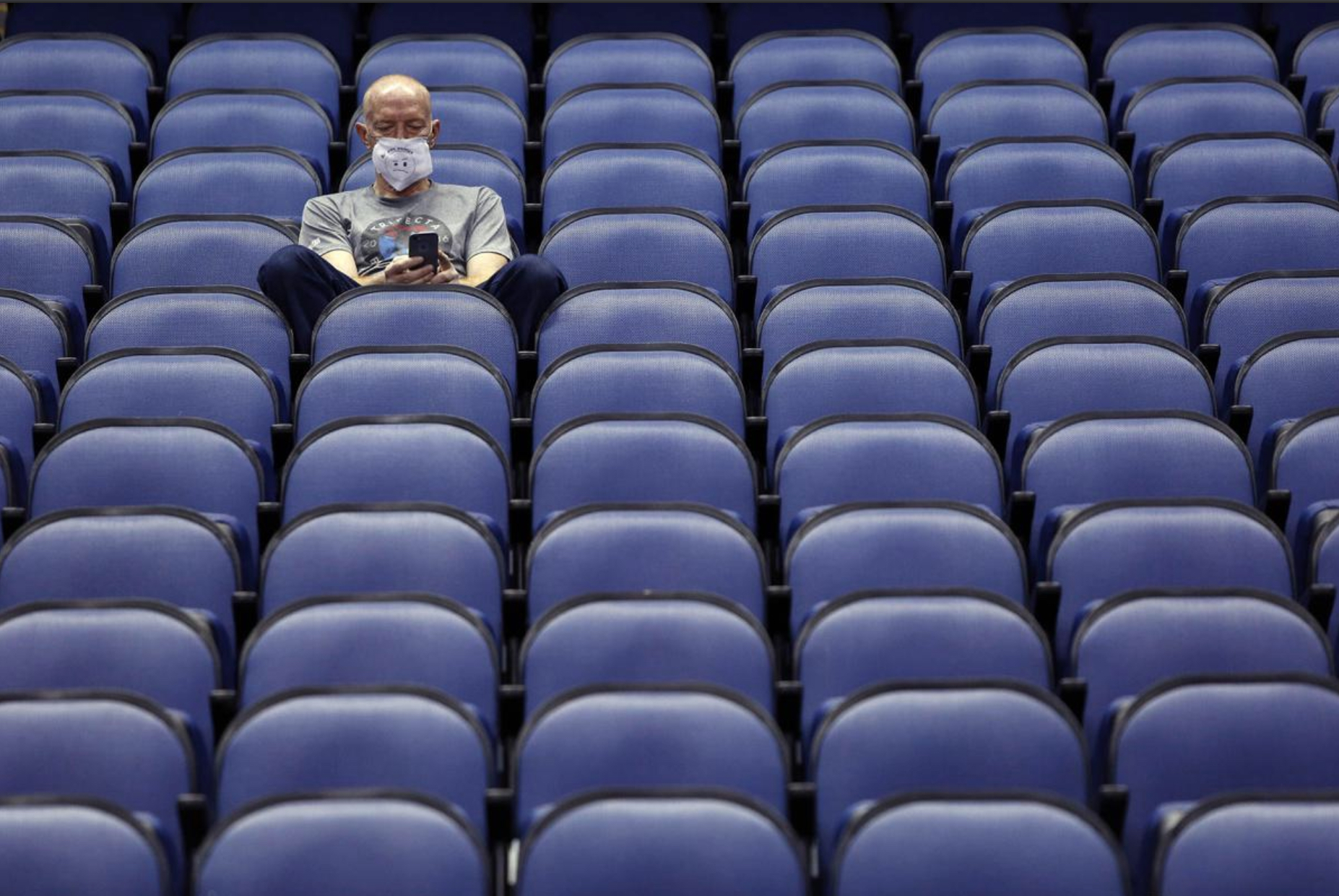 Photo shows a man wearing a surgical mask sitting alone in an empty stadium