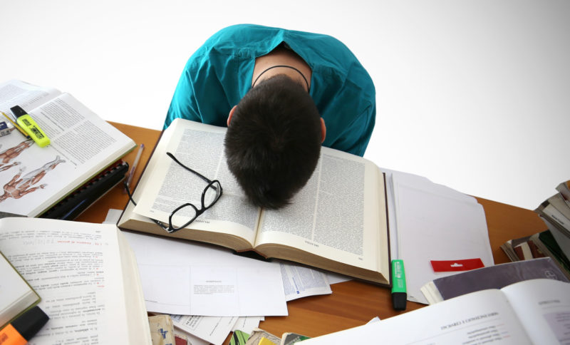 A student face palmed on his textbook and surrounded with many books and a glasses