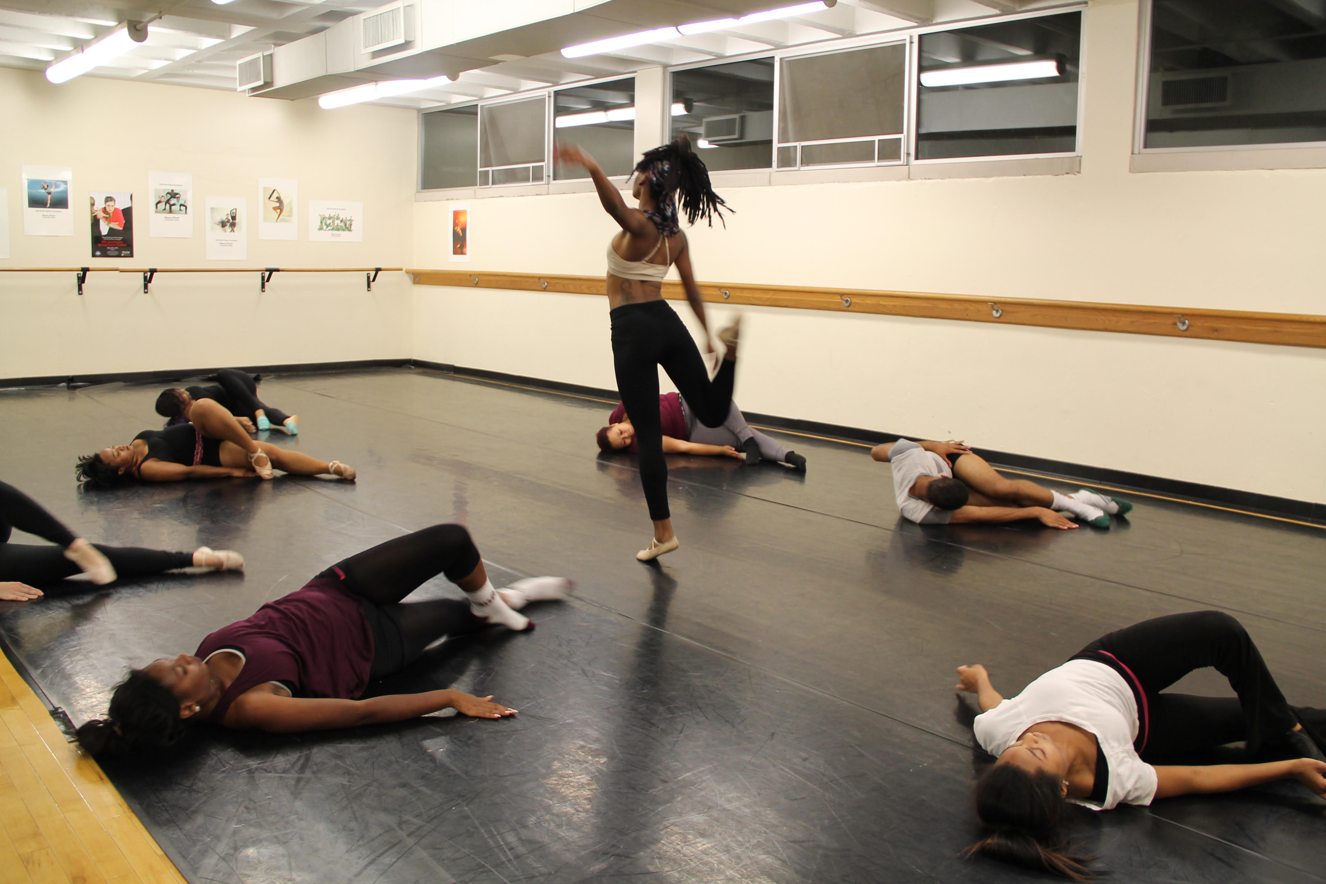 Dancers bending backward in circle around female dancer twirling on one leg in rehearsal studio.