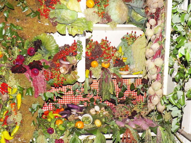 A cabinet overflows with fruit, and vegetables, leaves and moss. It has been arranged in an artful manner.