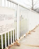 A handwritten sign reading "Refugees Welcome" is posted on a white fence that extends into the distance.