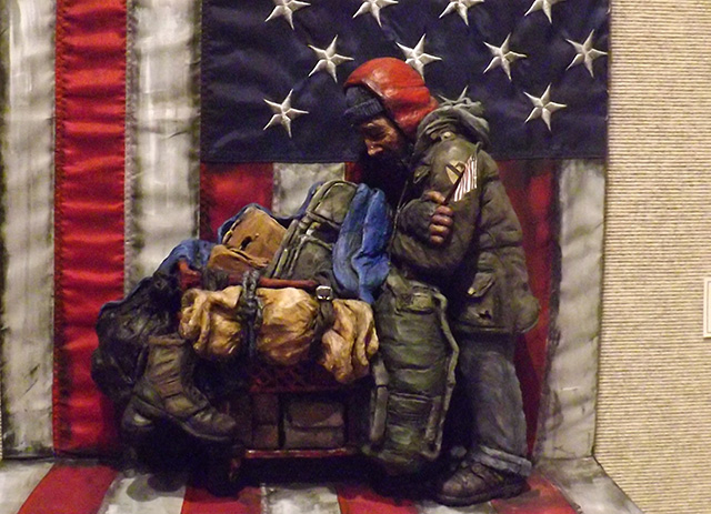 A ceramic sculpture of a man huddled by several bags set in a pile in a shopping cart. The man is in a large, puffy winter coat with a red winter cap. His arms are wrapped around himself as he gazes down at the pile. The background and base is that of an American flag.