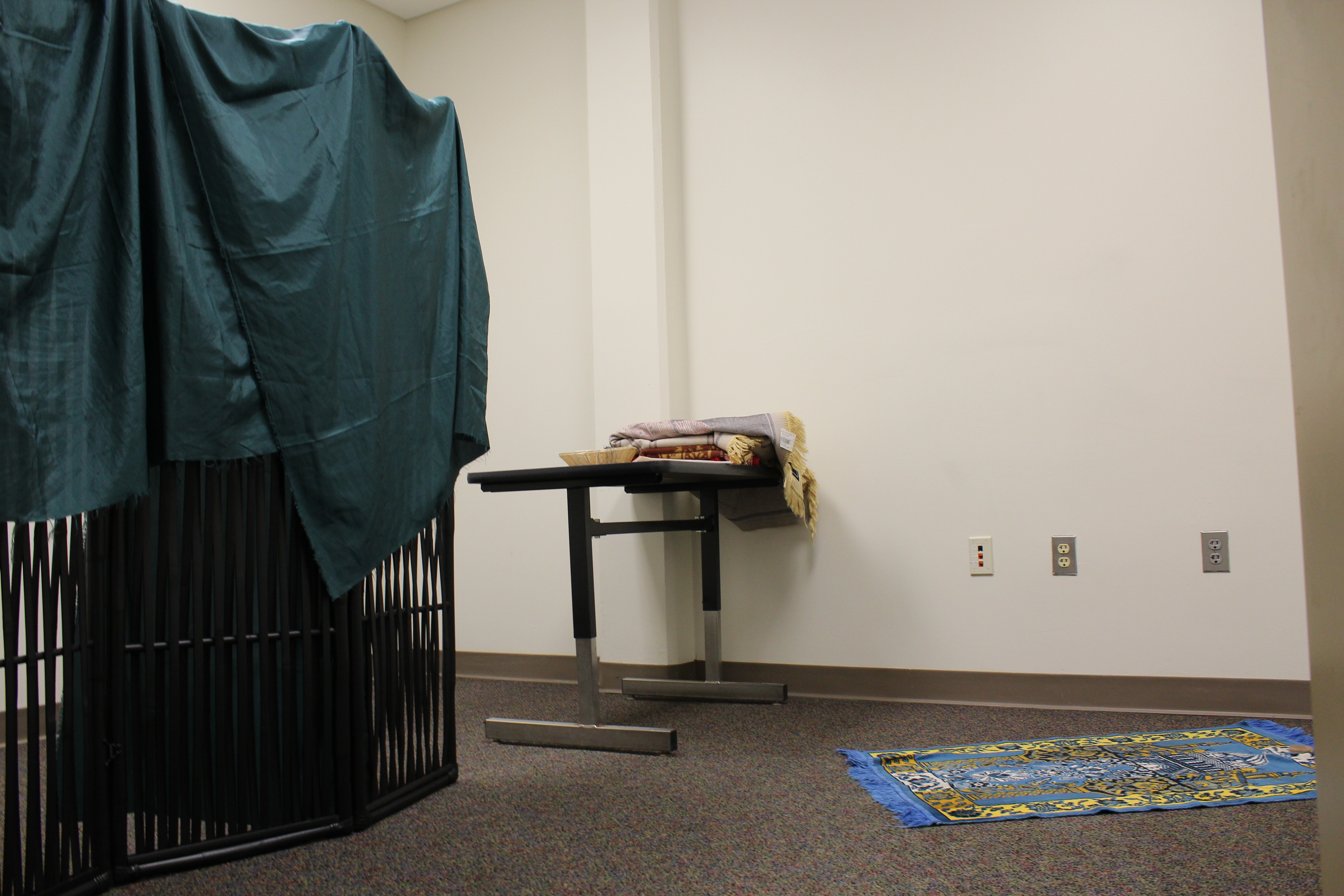 A simple room with a room divider draped with fabric on the left. A small table holds prayer mats and one is rolled out on the floor.