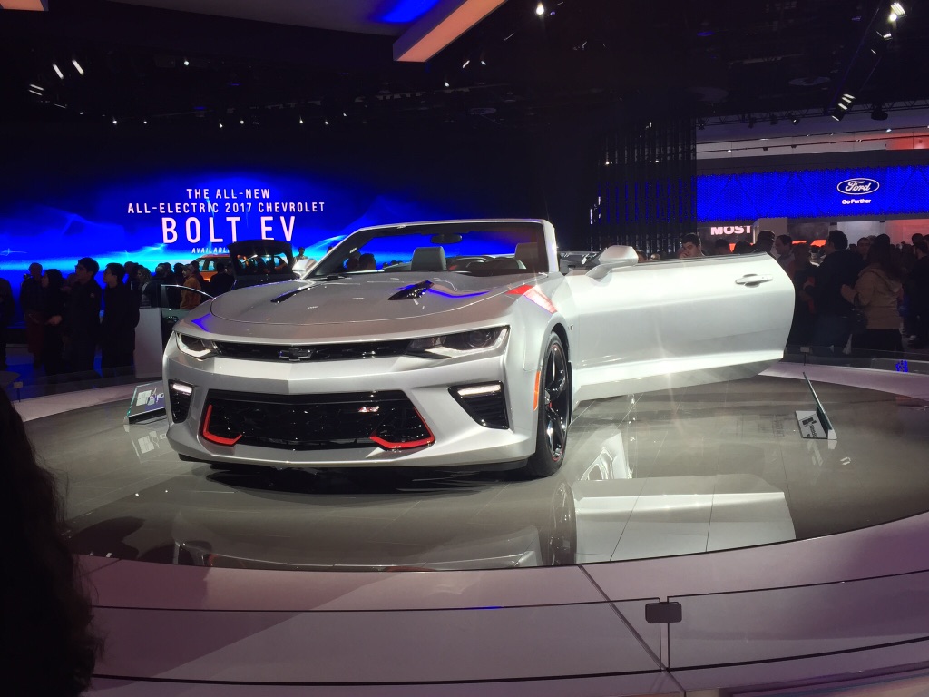 A new, shiny, silver convertible sit on a platform with the driver-side door opened.