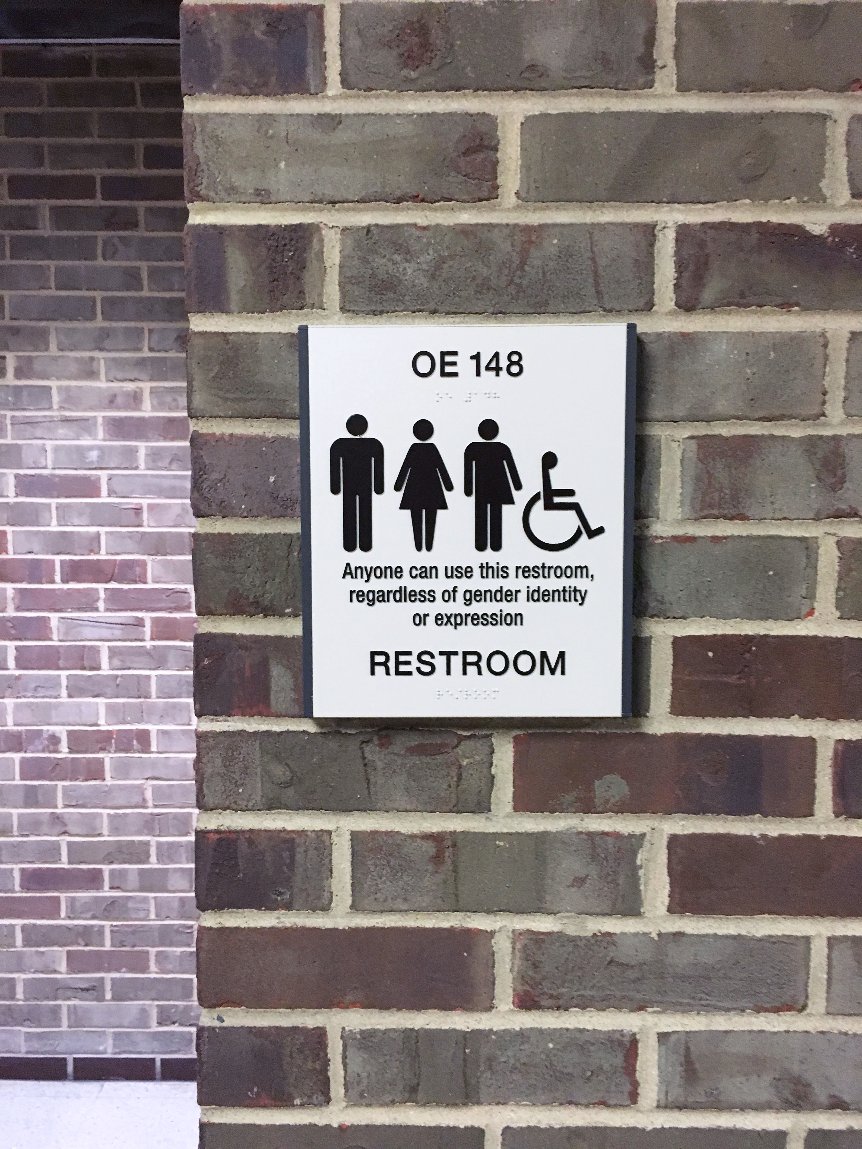 A brick wall displays a sign that has four symbols: a stick figure in pants typically denoting the male gender, a stick figure in a dress typically denoting the female gender, a stick figure that is wearing pants on one side and a dress on the other, and the stick figure denoting accessibility. The text on the sign notes it is a restroom and, "anyone can use this restroom, regardless of gender identity or expression".