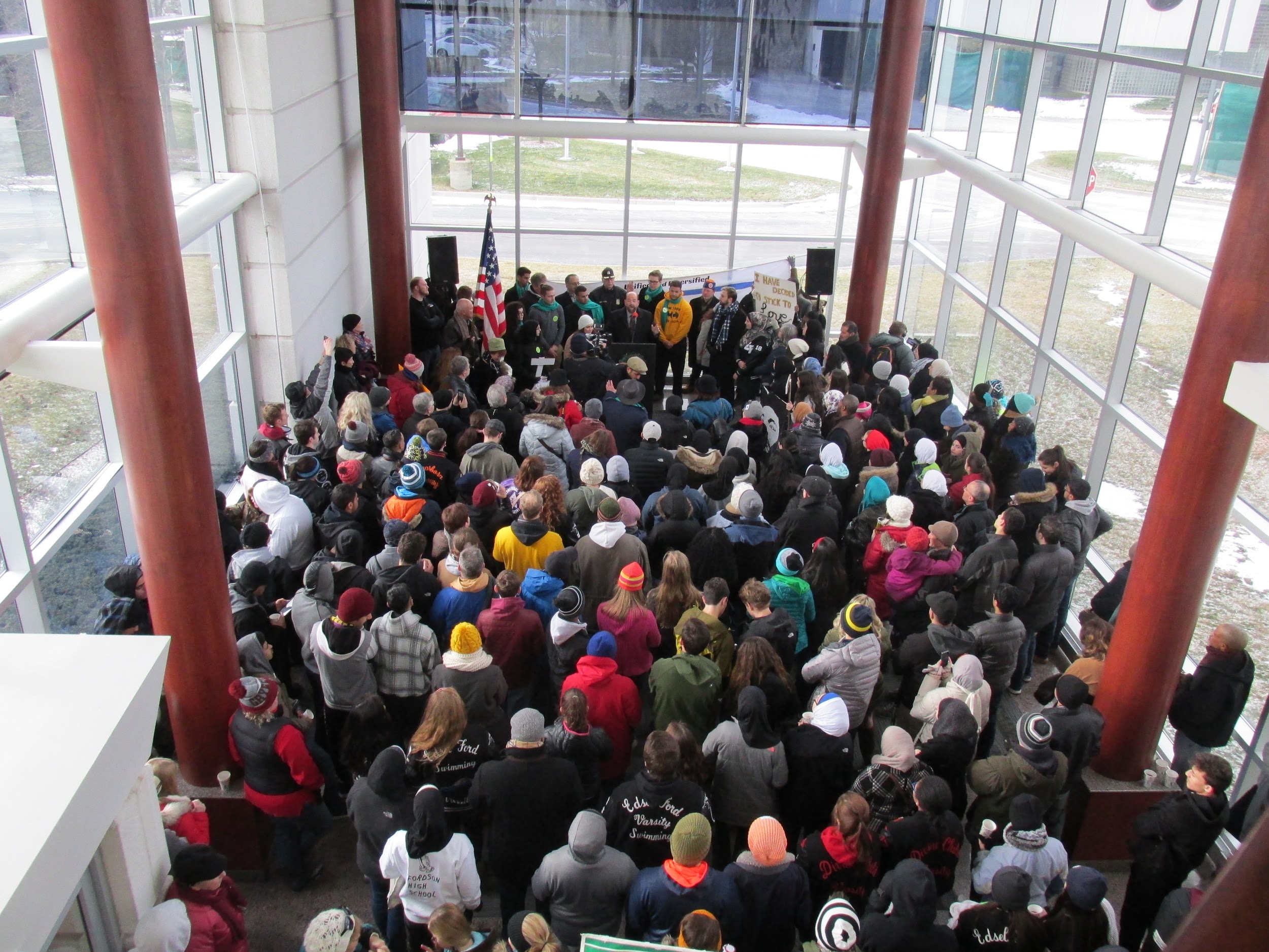 A top down view of roughly 100 people gathered in a lobby space. They stand in front of a podium where several people are standing.