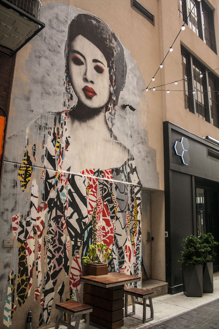 The brick exterior of a building has a large, detailed painting of an asian woman, who is depicted to be dressed in colorful strips, with elaborate patterns.