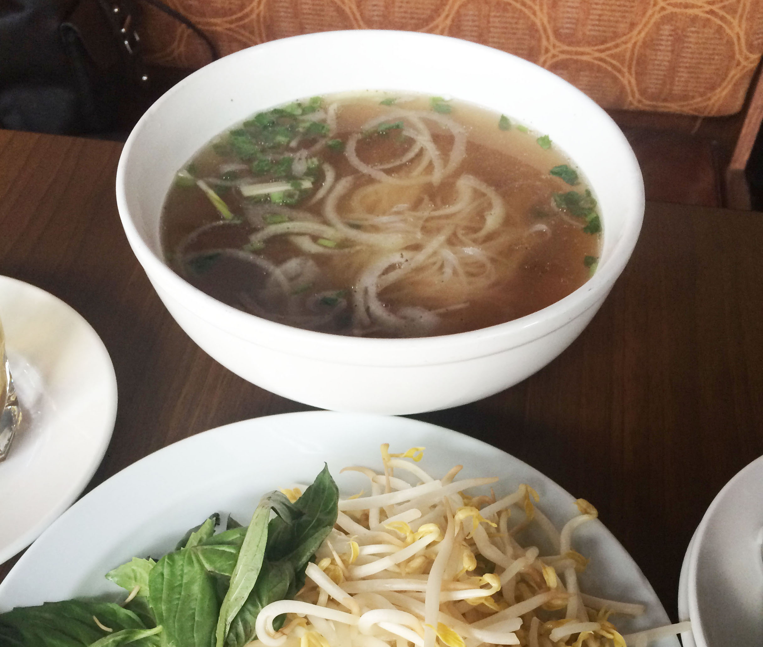 A large bowl of pho, a broth based Vietnamese soup, sit on a table filled with noodles. A plate next to this bowl holds bean sprouts and Thai basil.