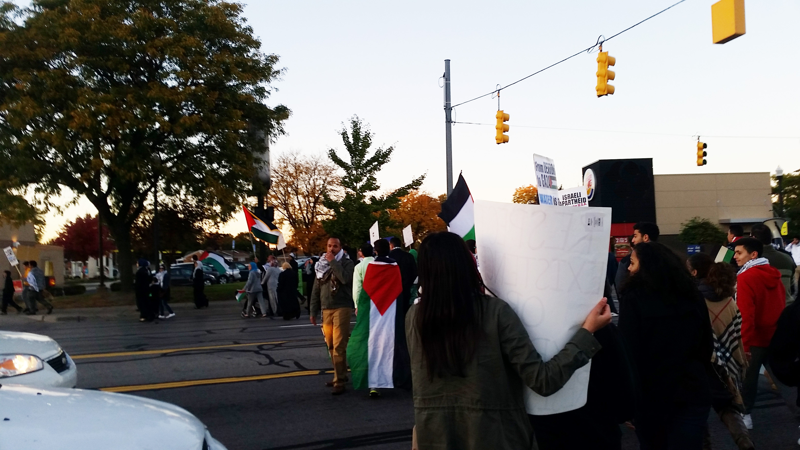 A row of protesters move across a street, holding signs and Palestinian flags.