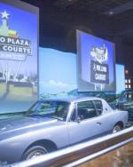 A classic car is displayed in front of photos of roadside signs. One sign reads, "Alamo Plaza Hotel Courts". It is in an art deco retro style.