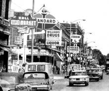 A black and white photo captures a busy street scene, with cars from the 1950s driving past a block filled with businesses with large signs. Some of the signs read: "Super Market" "Checks Cashed" "Standard [Oil]" "Williams Drugs"
