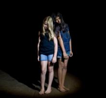 Two young girls in jean shorts and dark sleeveless shirts with long hair covering their faces in a dark backdrop.
