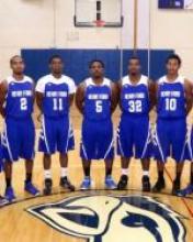 A team photo of the men's basketball team. The men stand in a line at center court in royal blue uniforms with white details.