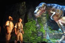 Two men in safari style gear gaze up at a rendered dinosaur. The dinosaur looks down at them.