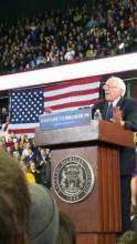 Bernie Sanders speaking from a podium at Eastern Michigan University. Behind him is a large American flag and many supporters.