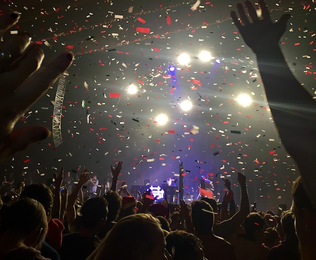 Confetti flies down from the ceiling inside a large concert hall. The crowd inside raises there hands up. Lights shine from the stage as a band plays.