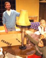 Actors Matt Van Houten, Brigid Driscoll, and Blues Higgerns sit in a staged living room and look on as Houten steadies a lamp. The lamp shade is yellow and of an older, dated style. It has a black fringe. The base of the lamp is a leg in fishnet stockings and a high-heeled shoe.