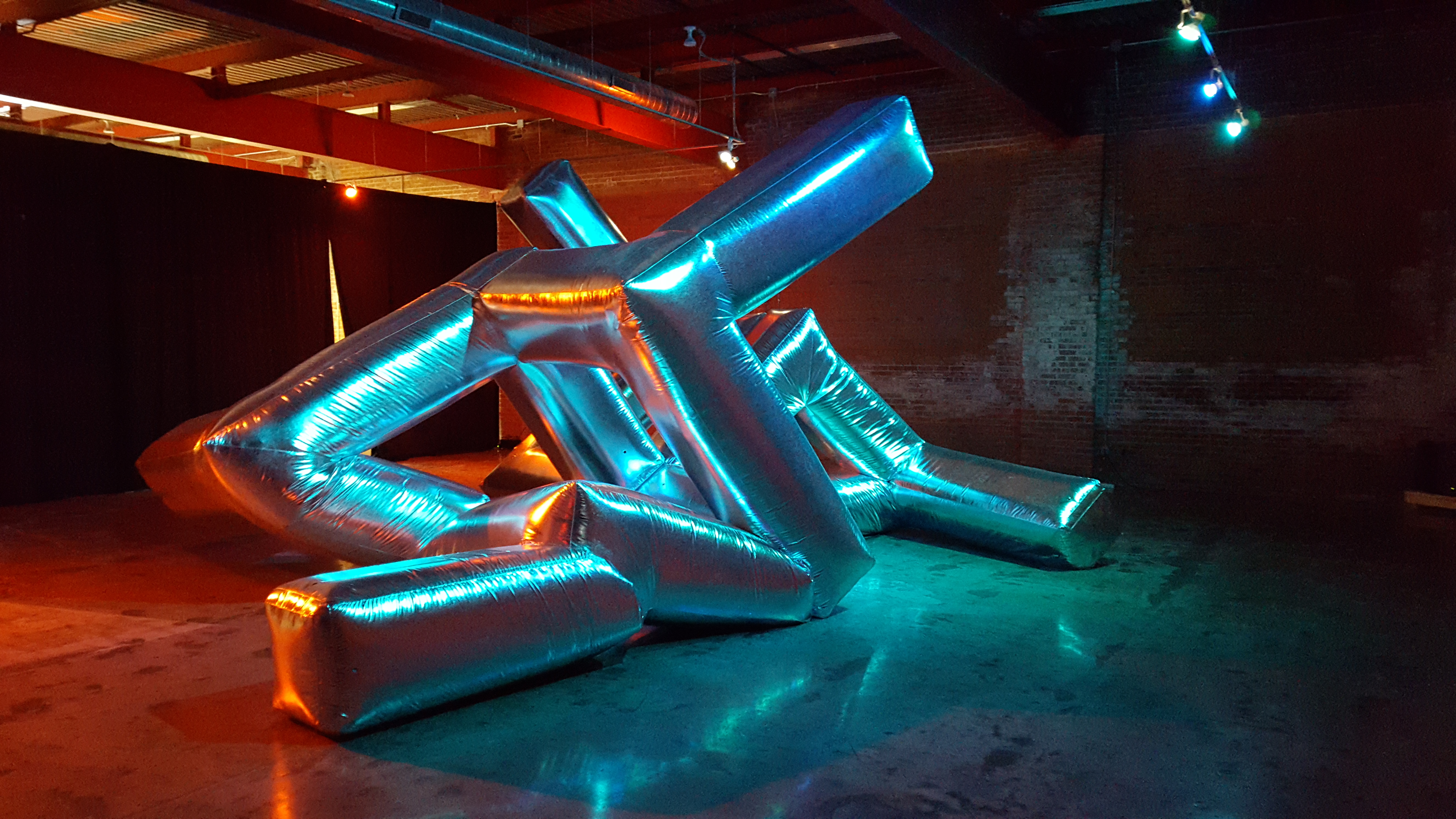 A shiny, inflated, metallic art piece fills a room. The "sculpture" is geometric, but appears to be stuffed or inflated. Several arms coming off of the piece, which bend at various points. It fills the large loft style space it is photographed in.